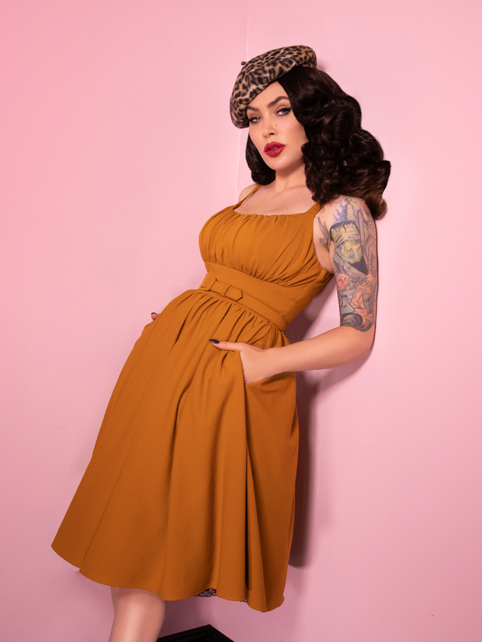 Micheline Pitt leaning against the wall wearing the vintage inspired Ingenue Dress in mustard print with her hands tucked into the pockets of the dress. 
