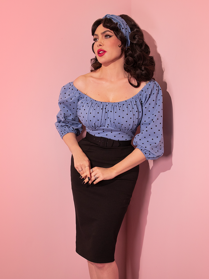 Micheline Pitt posing while wearing the Daydream Wiggle Dress in Sunset Blue Polka Dot tucked into a black pencil skirt.