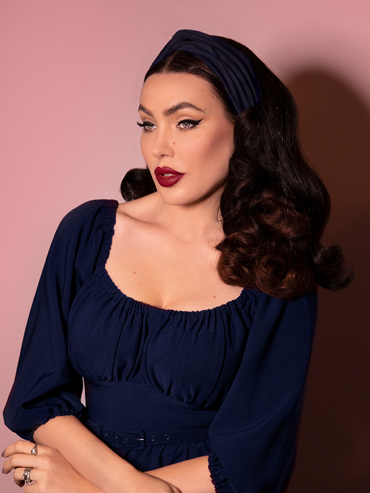 Looking off camera, Micheline Pitt wears a blue long sleeve top along with matching retro inspired knot headband in navy.