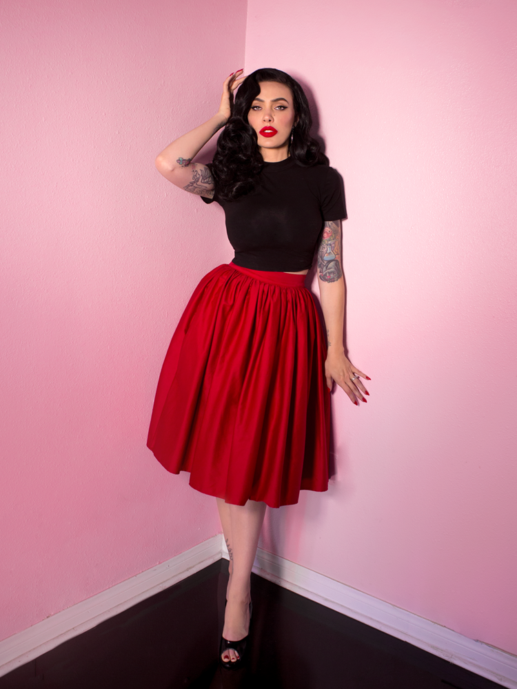 Full body length shot of Micheline Pitt standing against pinks walls wearing a classic retro outfit - Vixen Swing Skirt in Red and black top from Vixen Clothing.